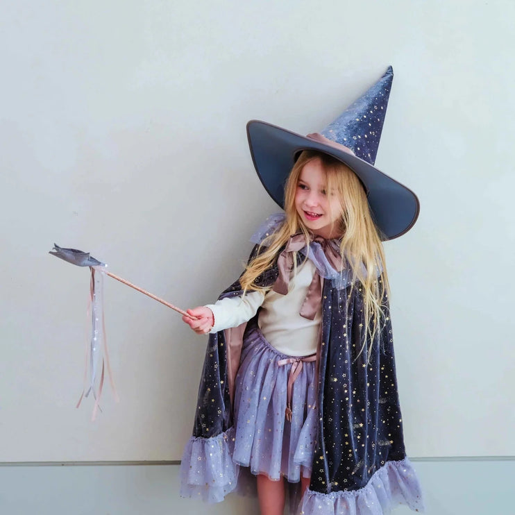 A young girl with long blonde hair wearing a wizard costume, including a dark blue starry hat, cape, and matching skirt, holding a wand against a plain light gray wall.