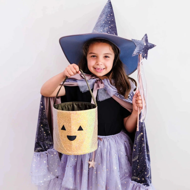 A young girl wearing a sparkly purple wizard costume with a pointed hat, cape, and skirt, holding a glittery gold jack-o'-lantern bucket and a star wand against a plain white background.