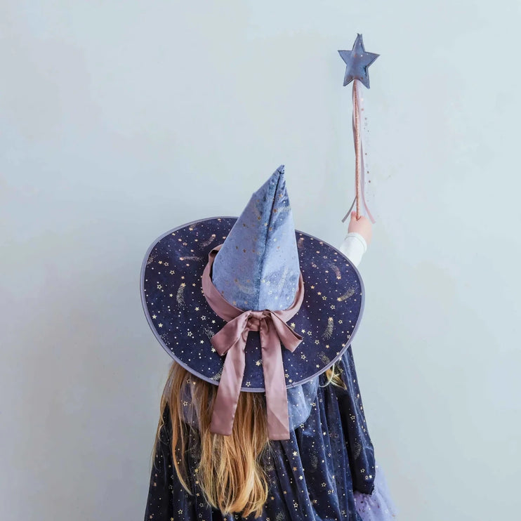 A child seen from behind wearing a wizard costume hat and matching starry cape, holding a star wand against a plain light wall.