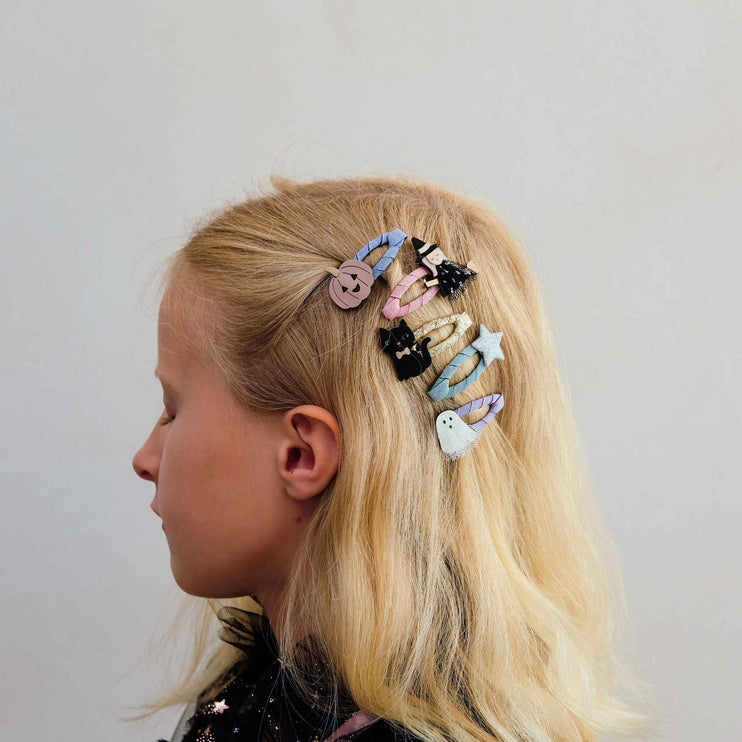 A side-profile view of a young girl with blonde hair wearing a set of colorful, whimsical hair clips, including a button, a ghost, a star, and a small cat