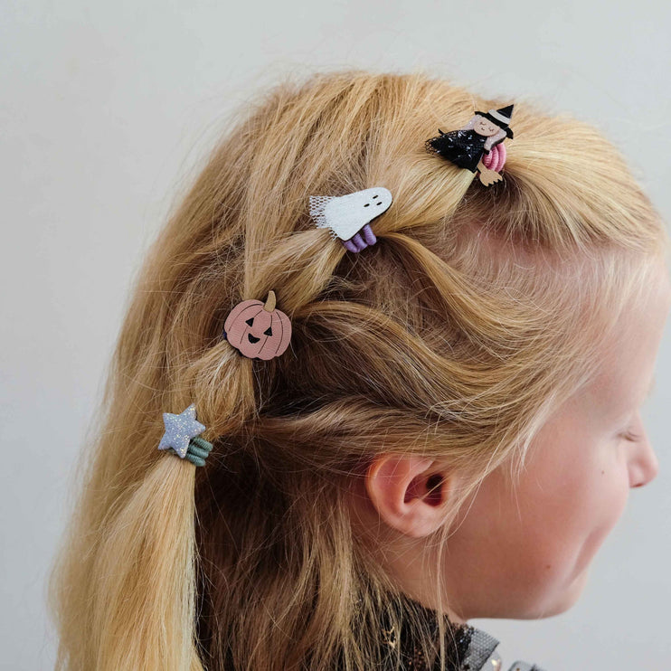 A close-up side profile of a young girl with blonde hair styled in a low ponytail, decorated with four small Halloween-themed hair clips including a jack-o'-lantern, ghost, witch, and star.