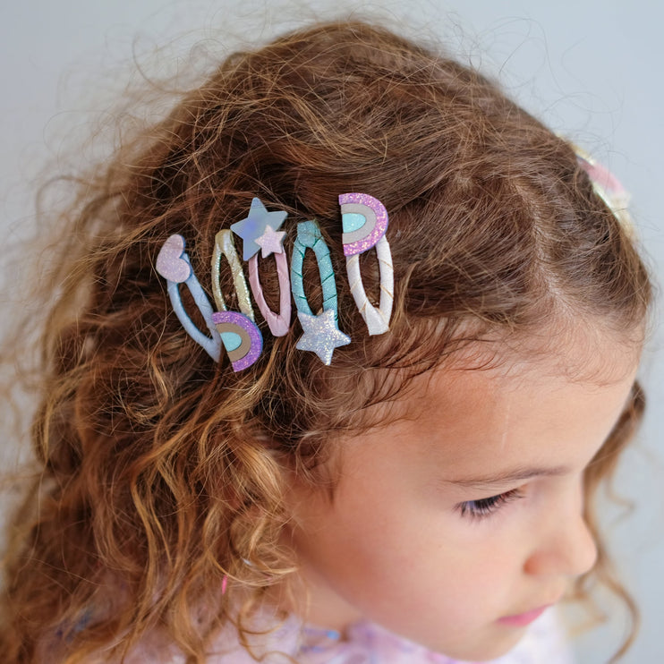 A close-up of a young girl with curly brown hair wearing a set of pastel rainbow and star hair clips.