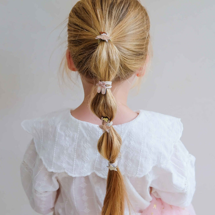 A child  wearing a white ruffled blouse, showing a blonde ponytail styled with three pastel hair ties 