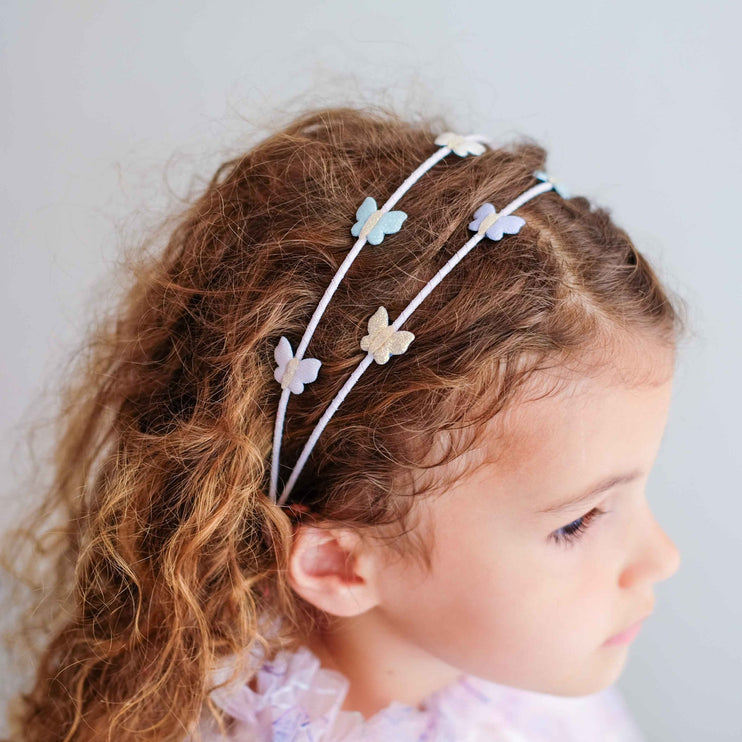 A young girl with curly brown hair wearing a white elastic headband decorated with small pastel butterfly charms
