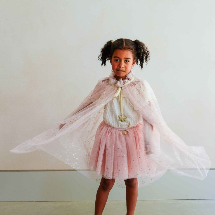 A young girl wearing a pink and white princess costume, featuring a sparkly pink tulle skirt, a white top with a gold star necklace, and a sheer white cape