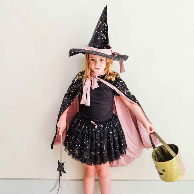 A young girl wearing a witch costume. The costume includes a black pointed hat with pink trim, a black tunic, a black sparkly skirt, and a pink cape.