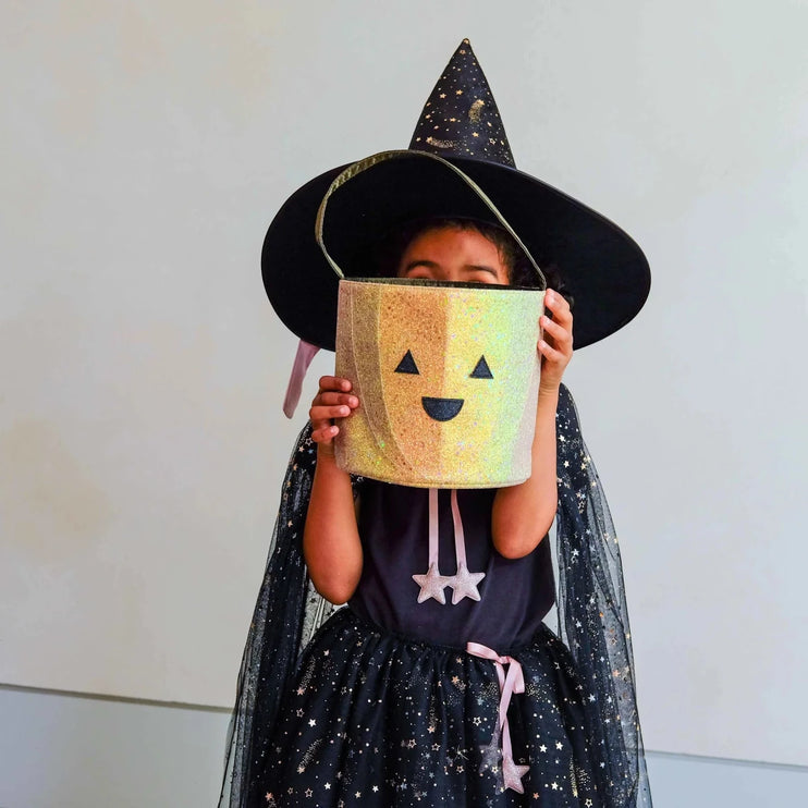 A child wearing a black witch hat with gold speckles, a black tulle cape with gold stars, and a dark dress holds a glittery gold jack-o'-lantern bucket with a black face on a plain light background.