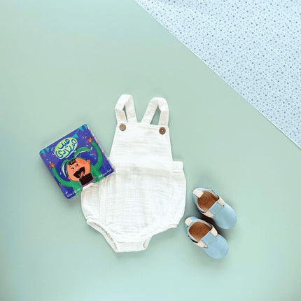 White baby romper with suspenders, blue leather shoes, and a children's book 
