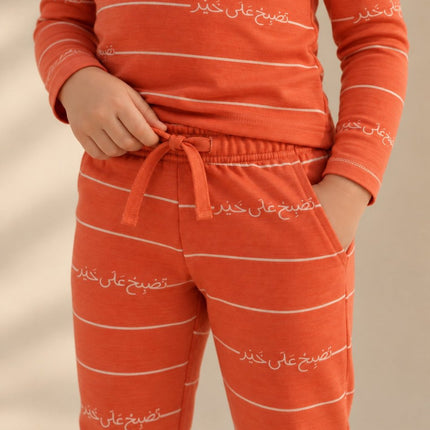 A child wearing a matching orange long-sleeve top and jogger pants set with thin white horizontal stripes and white Arabic script printed