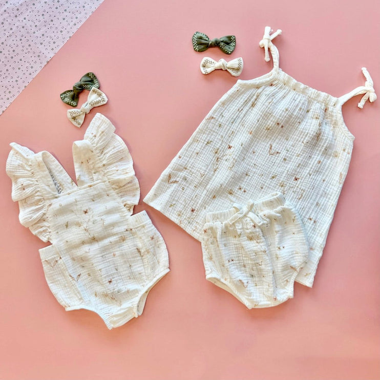Baby romper set with ruffled top and bloomers, accompanied by two hair bows on a pink background.