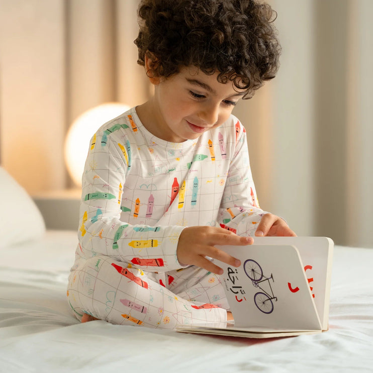 Child wearing Alwan pajama with colorful crayon illustrations and text. reading a book on a bed.