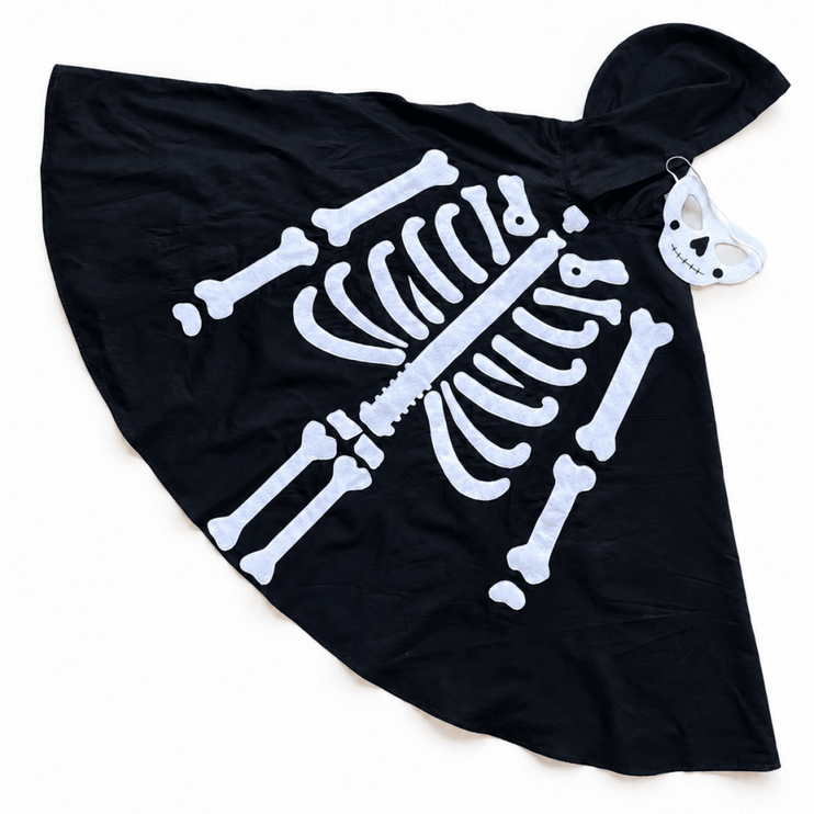A black hooded cape or poncho laid flat on a white background, featuring a large white skeleton bone print across the front. A small white skull-and-bone tag is attached near the hood.