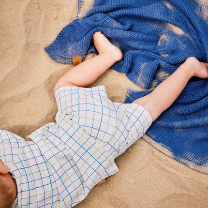 Baby boy wearing cream top and pants with blue and green stripes lying on beach sand
