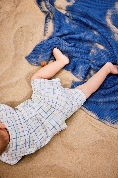 Baby boy wearing cream top and pants with blue and green stripes lying on beach sand