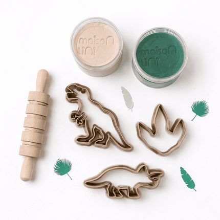 Cookie cutters with dinosaur designs, a rolling pin, and two containers of dough on a white background.