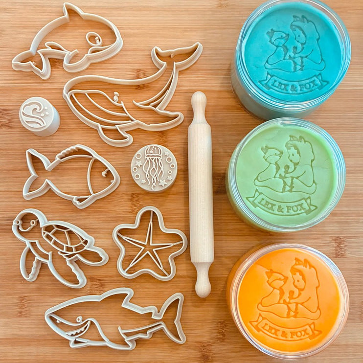 A set of sea-themed cutters and stamp molds  with three round tubs of modeling clay in turquoise, mint green, and orange. A small wooden rolling pin is also included