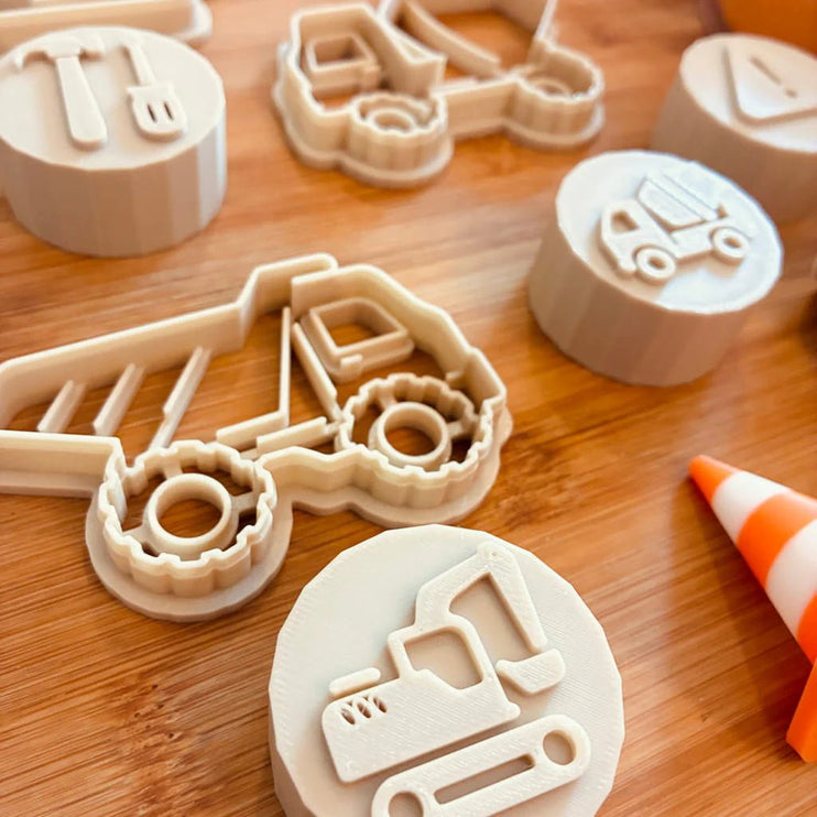 A set of 3D-printed construction-themed cookie cutters and stamps in a light beige plastic, displayed on a wooden surface. The set includes various shapes like a dump truck, excavator, crane, and traffic cones.