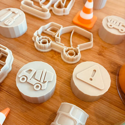 A set of light beige plastic toy construction and traffic-themed cookie cutters and stamps arranged on a wooden surface, including a truck, dump truck, warning sign, and traffic cone designs.