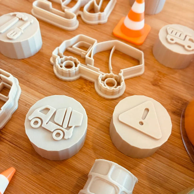 A set of light beige plastic toy construction and traffic-themed cookie cutters and stamps arranged on a wooden surface, including a truck, dump truck, warning sign, and traffic cone designs.