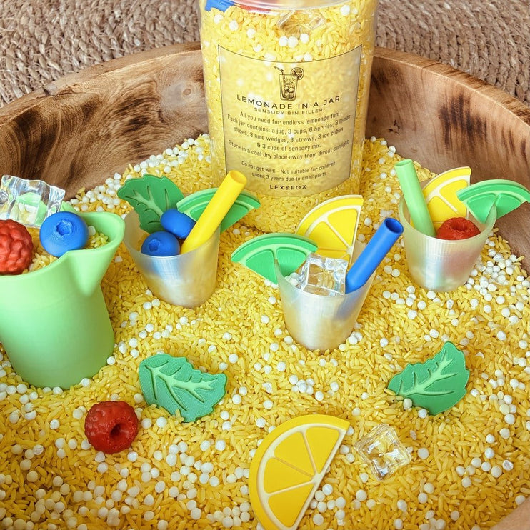 A colorful DIY craft kit for making 'Lemonade in a Jar' displayed in a wooden bowl filled with yellow beads. The kit includes a clear bottle with a yellow label, miniature metal cups with blue and yellow straws, green and yellow lemon slices, raspberries, a green pitcher, and green leaf-shaped beads.