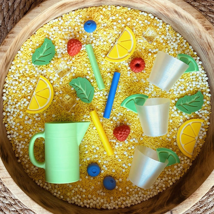 A sensory play bin filled with yellow beads, featuring plastic toy cups, straws, lemon slices, berries, and a green watering can.