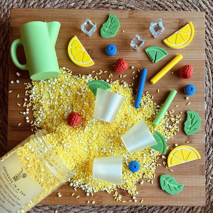 A colorful lemon-themed sensory play set arranged on a wooden board, including a green pitcher, yellow sprinkles, white and yellow beads, lemon slices, blueberries, green leaves, and small plastic cups. 