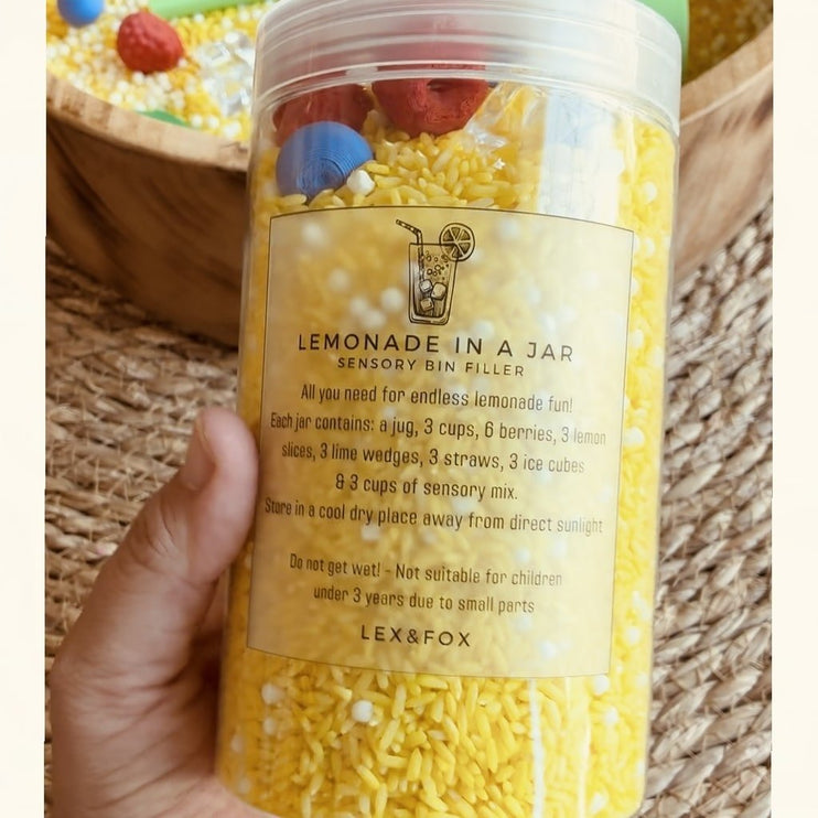 A hand holding a clear plastic jar filled with yellow and white textured sensory bin filler labeled 'LEMONADE IN A JAR' by Lex&Fox