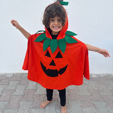 A child wearing a bright orange pumpkin costume cape with green leaves and a black jack-o'-lantern face, standing barefoot on a paved surface against a white wall.