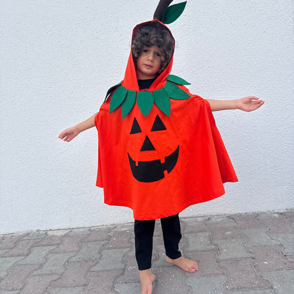 A child wearing a bright orange pumpkin costume cape with a black jack-o'-lantern face, green leafy collar detail, and a black stem on the hood, standing barefoot outdoors against a white wall.