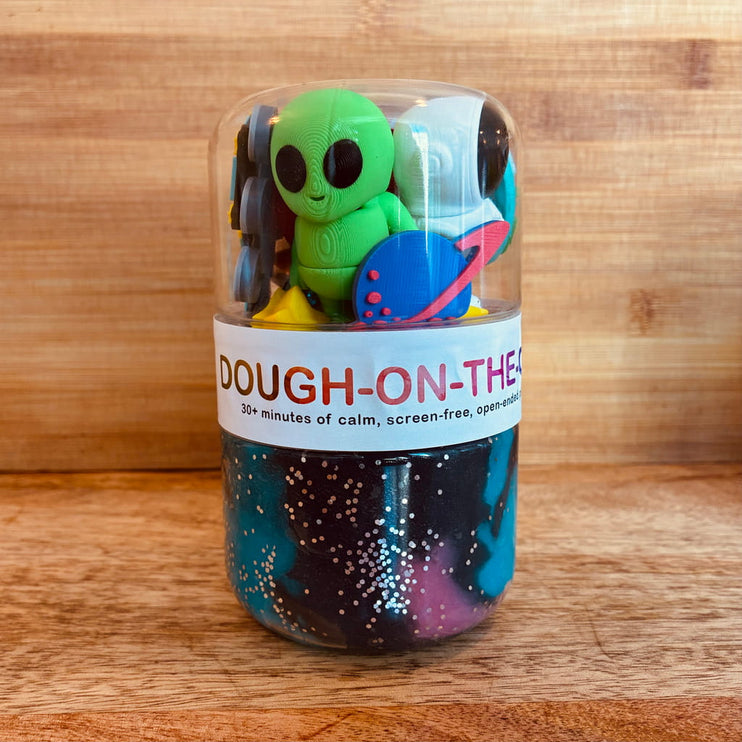 Toy set with alien figure and planet in a clear container on a wooden surface, branded 'DOUGH-ON-THE'.