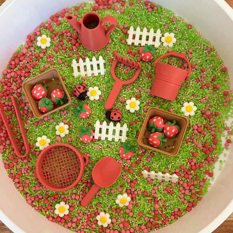 Sensory bin with green and red play materials including a watering can, flowers, and small boxes.