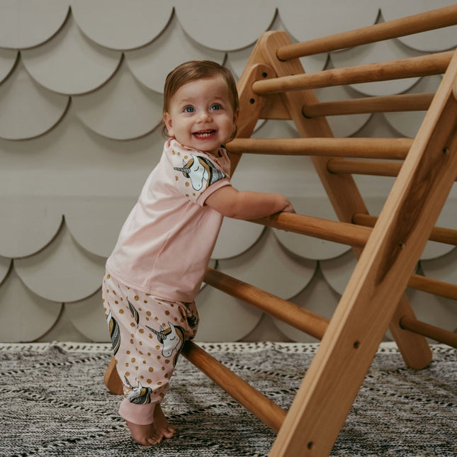 Child in pink outfit standing next to a wooden climbing toy against a scalloped wall.