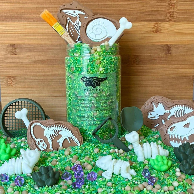A dinosaur-themed sensory bin. The set includes dinosaur skeleton and skull cutouts on brown shapes, white bones, green rocks, purple rhinestones, small black toy tools, and a paintbrush