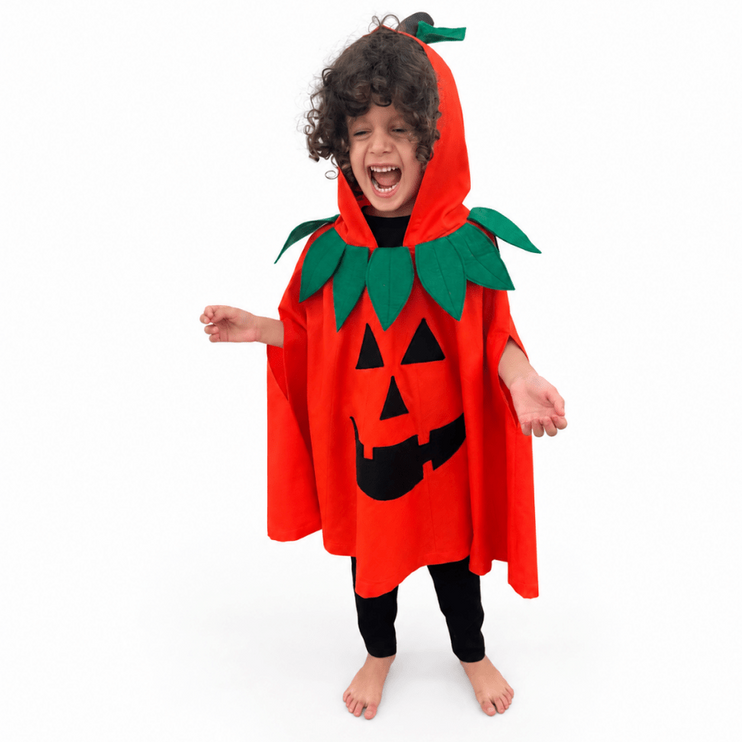 A child wearing a bright red pumpkin costume cape with a black jack-o'-lantern face and green leafy collar details, standing barefoot against a plain white studio background.