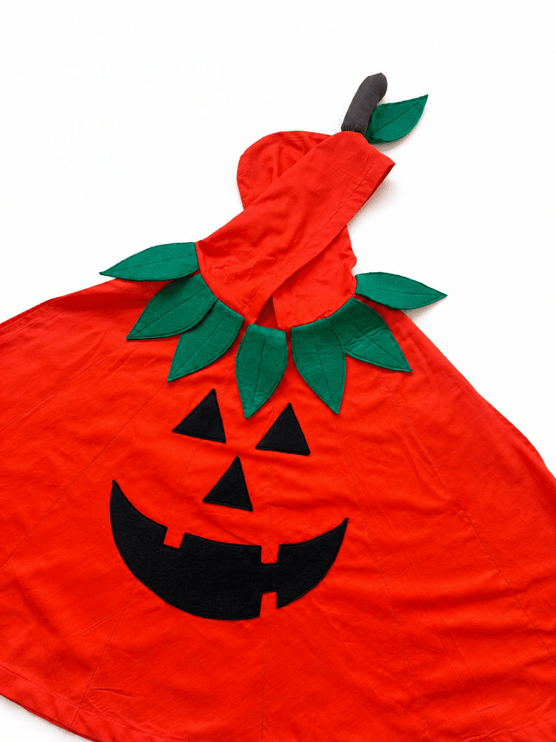 A bright red Halloween pumpkin costume cape with a black embroidered jack-o'-lantern face, green felt leaves, and a small dark stem on a white background.