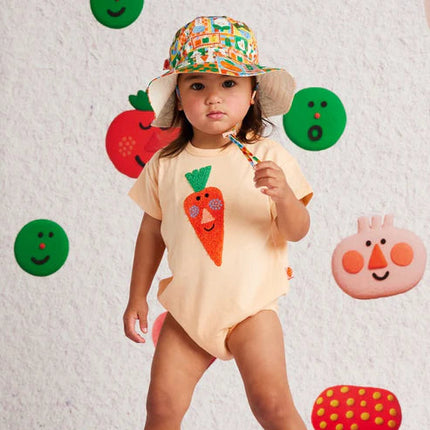 Child wearing a onesie with a carrot design and a colorful hat, standing against a wall with fruit and vegetable decorations.