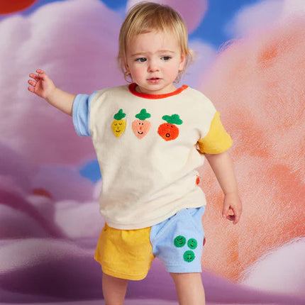 Child wearing a colorful outfit with fruit designs against a soft, multicolored background